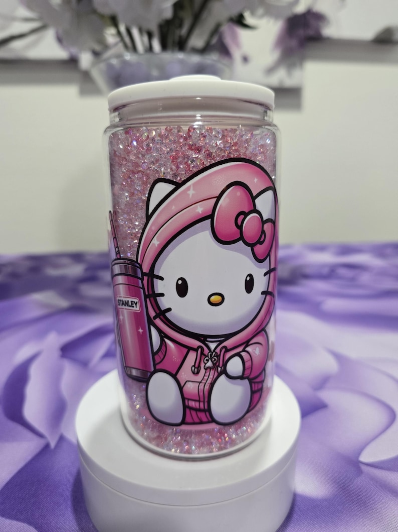 May include: A clear, cylindrical tumbler filled with pink glitter and a cartoon graphic of Hello Kitty in a pink hoodie. A pink Stanley tumbler graphic is also visible. The tumbler has a white lid and sits on a white stand.