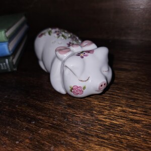 May include: A white ceramic pig figurine with pink ears and a pink bow. The pig is painted with pink and purple flowers and has closed eyes. The pig is lying on its side.
