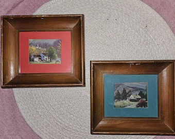 May include: Two framed landscape paintings with wooden frames. The painting on the left has a red mat and depicts a small village with a blue roofed house. The painting on the right has a green mat and depicts a small village with a white roofed house.