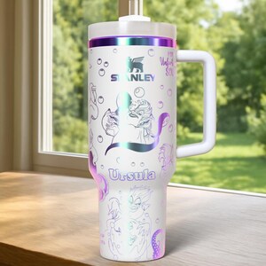 Może przedstawiać: Biały kubek Stanley z uchwytem, z motywem Urszuli z Małej Syrenki. Kubek ma białą pokrywkę i uchwyt, z iryzującymi akcentami. Widoczny jest napis "Urszula" i "For Unfortunate Souls".