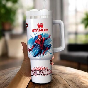 Op de afbeelding: Witte Stanley-beker met een Spider-Man-ontwerp. De beker heeft een handvat, een rietje en het Stanley-logo. De onderkant van de beker leest "MARVEL ULTIMATE SPIDER-MAN". De beker is ontworpen om een grote hoeveelheid vloeistof te bevatten.