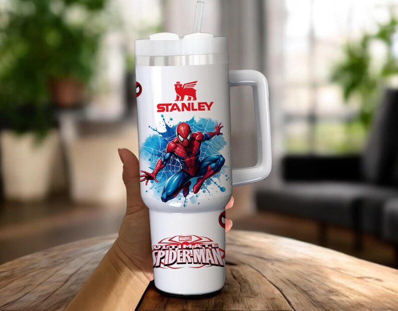 Op de afbeelding: Witte Stanley-beker met een Spider-Man-ontwerp. De beker heeft een handvat, een rietje en het Stanley-logo. De onderkant van de beker leest "MARVEL ULTIMATE SPIDER-MAN". De beker is ontworpen om een grote hoeveelheid vloeistof te bevatten.