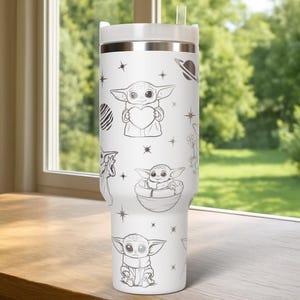 May include: White stainless steel tumbler with a silver lid and straw. The tumbler is decorated with black and white illustrations of a character holding a heart, in a pod, and other space-themed designs. The tumbler is sitting on a wooden surface.