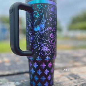 Puede incluir: Vaso t&eacute;rmico negro con asa, con un dise&ntilde;o colorido de un personaje de dibujos animados tocando el ukelele, estrellas y flores. El vaso tiene una tapa y pajita negras, con un degradado de tonos azules y morados.