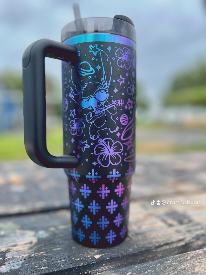 Puede incluir: Vaso t&eacute;rmico negro con asa, con un dise&ntilde;o colorido de un personaje de dibujos animados tocando el ukelele, estrellas y flores. El vaso tiene una tapa y pajita negras, con un degradado de tonos azules y morados.