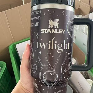 May include: A black and silver Stanley travel mug with a black handle. The mug features the word "Twilight" in white script, along with other white graphics and text. The mug has a black lid and is held by a person.