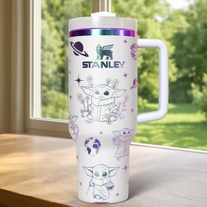May include: White Stanley tumbler with a handle, featuring illustrations of a character and space-themed graphics. The tumbler has a white lid and a purple and green iridescent band. The Stanley logo is visible on the front.