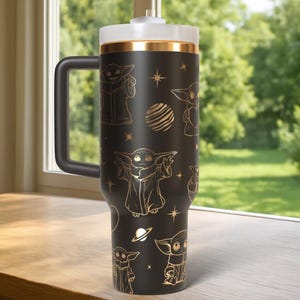 May include: A black travel mug featuring gold illustrations of a character, stars, and planets. The mug has a black handle, a white lid, and a gold band. The mug is designed to hold a large quantity of liquid.