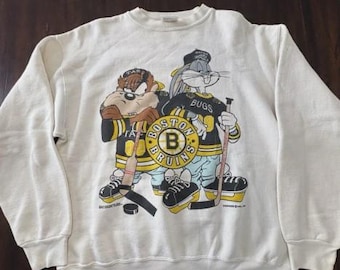 May include: Off-white sweatshirt featuring Taz and Bugs Bunny in Boston Bruins hockey jerseys. The graphic includes the team logo and hockey sticks. The sweatshirt has a crew neck and long sleeves.