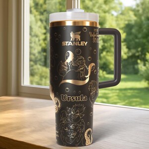 Może przedstawiać: Czarny kubek Stanley ze złotymi wzorami Ursuli i stworzeń morskich. Posiada białą pokrywkę, złote wykończenia i czarny uchwyt. Kubek ma logo Stanley i napis "Poor Unfortunate Souls".