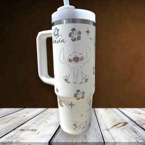 Może przedstawiać: Biały kubek termiczny z uchwytem, ​​z motywem Stitcha z Lilo & Stitch, wraz z kwiatami hibiskusa i słowem "Hawaii". Zawiera białą pokrywkę i słomkę. Kubek ma około 25 cm wysokości.