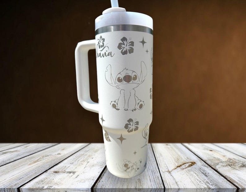 Może przedstawiać: Biały kubek termiczny z uchwytem, ​​z motywem Stitcha z Lilo & Stitch, wraz z kwiatami hibiskusa i słowem "Hawaii". Zawiera białą pokrywkę i słomkę. Kubek ma około 25 cm wysokości.