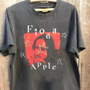Peut inclure: Un t-shirt noir &agrave; manches courtes avec un graphique rectangulaire rouge sur la poitrine. Le graphique affiche le portrait d'une personne avec le texte Fiona Apple imprim&eacute; en blanc, entour&eacute; de plusieurs &eacute;toiles blanches dessin&eacute;es &agrave; la main.