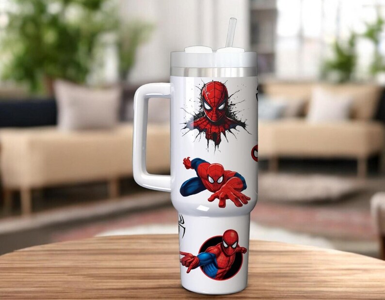 Op de afbeelding: Een witte beker met een handvat en een rietje, versierd met rode en blauwe Spider-Man afbeeldingen. De beker heeft een wit deksel en een grote handgreep. Het ontwerp toont verschillende actieposes van de superheld.