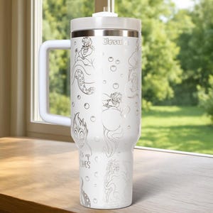 Może przedstawiać: Biały kubek termiczny z uchwytem, ​​z srebrnymi ilustracjami Urszuli z Małej Syrenki. Kubek ma białą pokrywkę i uchwyt, z napisem "Ursula" na g&oacute;rze. Widoczny jest r&oacute;wnież tekst "It's up to witches".