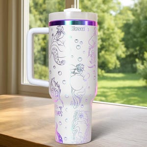 Może przedstawiać: Biały kubek termiczny z uchwytem, z Urszulą z Małej Syrenki. Projekt zawiera ilustracje liniowe Urszuli i tekst "It's up to witches". Kubek ma białą pokrywę i fioletowy, opalizujący pasek.