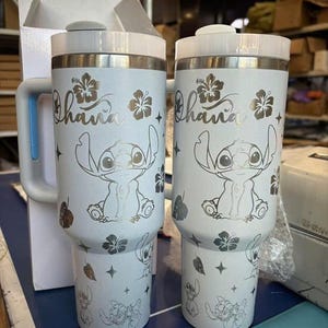 Może przedstawiać: Dwa białe kubki termiczne z uchwytem, z motywem Stitcha z Lilo & Stitch. Kubki mają napis "Ohana" i kwiatowe akcenty. Kubki są przeznaczone do utrzymywania zimnych lub gorących napoj&oacute;w.