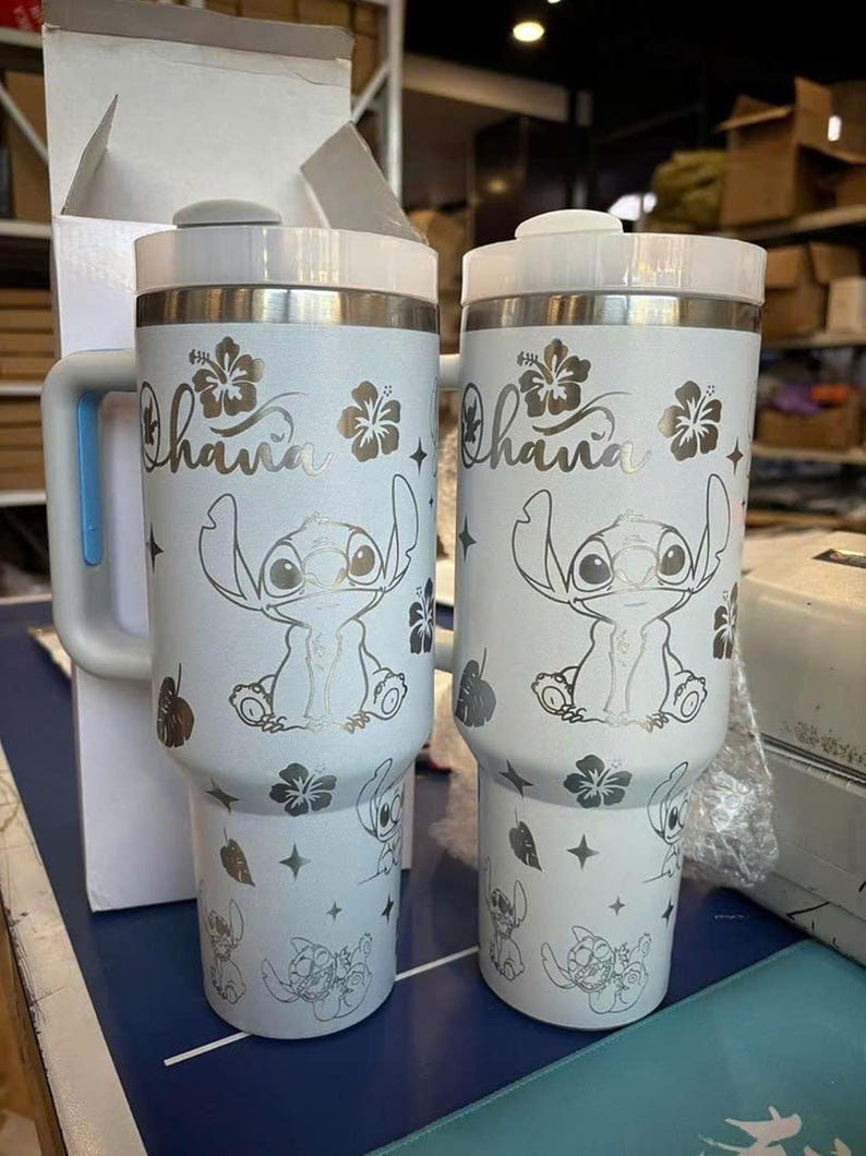 Może przedstawiać: Dwa białe kubki termiczne z uchwytem, z motywem Stitcha z Lilo & Stitch. Kubki mają napis "Ohana" i kwiatowe akcenty. Kubki są przeznaczone do utrzymywania zimnych lub gorących napoj&oacute;w.