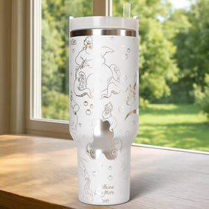 Może przedstawiać: Biały, izolowany kubek z srebrnym obrysem Urszuli z Małej Syrenki, bąbelkami i napisem "Villains Have More Fun". Kubek ma białą pokrywkę i przezroczystą słomkę. Kubek stoi na drewnianej powierzchni.