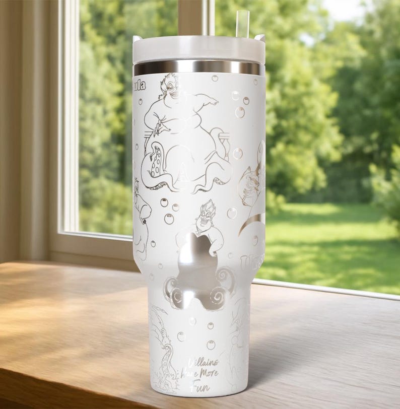 Może przedstawiać: Biały, izolowany kubek z srebrnym obrysem Urszuli z Małej Syrenki, bąbelkami i napisem "Villains Have More Fun". Kubek ma białą pokrywkę i przezroczystą słomkę. Kubek stoi na drewnianej powierzchni.