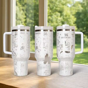 Może przedstawiać: Trzy białe kubki termiczne ze srebrnymi ilustracjami Urszuli z Małej Syrenki. Każdy kubek ma biały uchwyt, pokrywkę i słomkę. Kubki są ozdobione wizerunkiem Urszuli i napisem "Urszula". Jeden kubek ma logo Stanley.