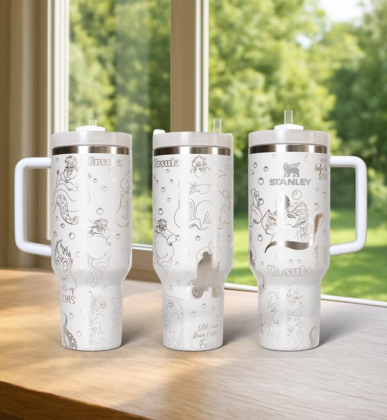 Może przedstawiać: Trzy białe kubki termiczne ze srebrnymi ilustracjami Urszuli z Małej Syrenki. Każdy kubek ma biały uchwyt, pokrywkę i słomkę. Kubki są ozdobione wizerunkiem Urszuli i napisem "Urszula". Jeden kubek ma logo Stanley.