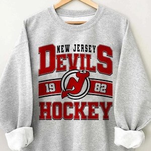 Peut inclure: Sweat-shirt gris avec un logo de hockey des Devils du New Jersey en rouge, blanc et noir. Le logo comprend le texte "New Jersey Devils" et les années "19" et "82". Le texte "Hockey" est sous le logo.