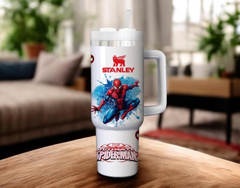 Op de afbeelding: Witte Stanley-beker met een Spider-Man-afbeelding. De beker heeft een handvat, een wit deksel en een rietje. De Spider-Man-afbeelding toont de superheld in een dynamische pose met blauwe en rode kleuren. De beker heeft ook de tekst "ULTIMATE SPIDER-MAN".