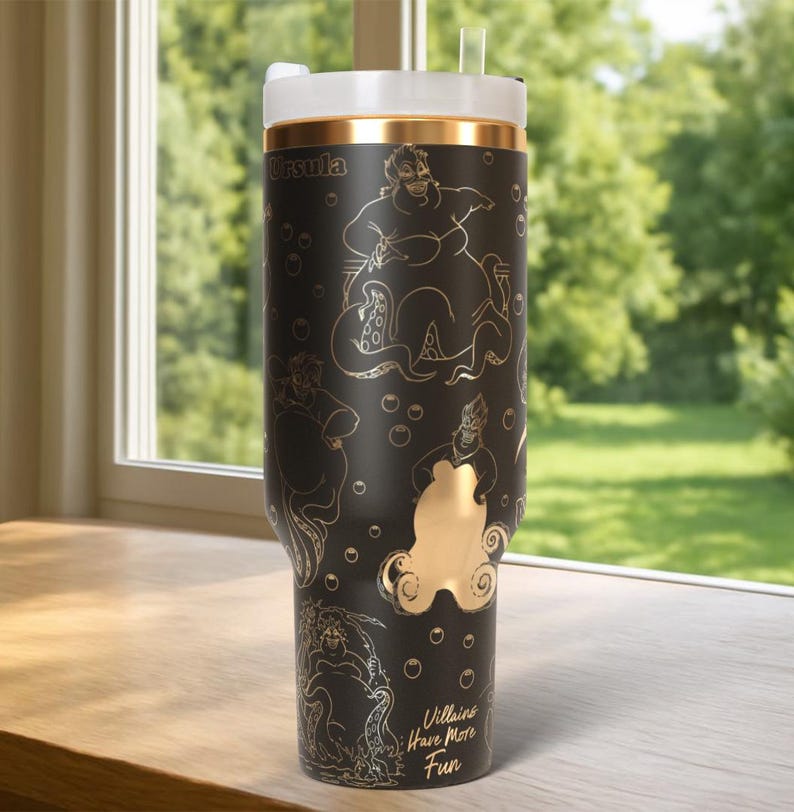 Może przedstawiać: Czarny kubek ze złotymi akcentami i białą pokrywką. Kubek zawiera złote ilustracje Urszuli z Małej Syrenki, bąbelki i napis "Villains Have More Fun."