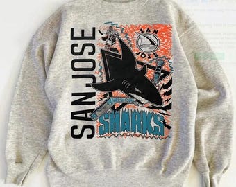 May include: A light gray sweatshirt featuring a graphic of a black shark with hockey sticks and the words "SAN JOSE SHARKS" in black and teal. The design includes orange and teal accents, and the sweatshirt has a crew neck and long sleeves.