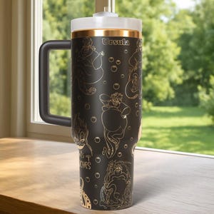 Może przedstawiać: Czarny kubek podr&oacute;żny z białą pokrywką i czarnym uchwytem. Kubek zawiera złote ilustracje Urszuli z Małej Syrenki, z widocznym napisem "Urszula" i frazą "Witches up".