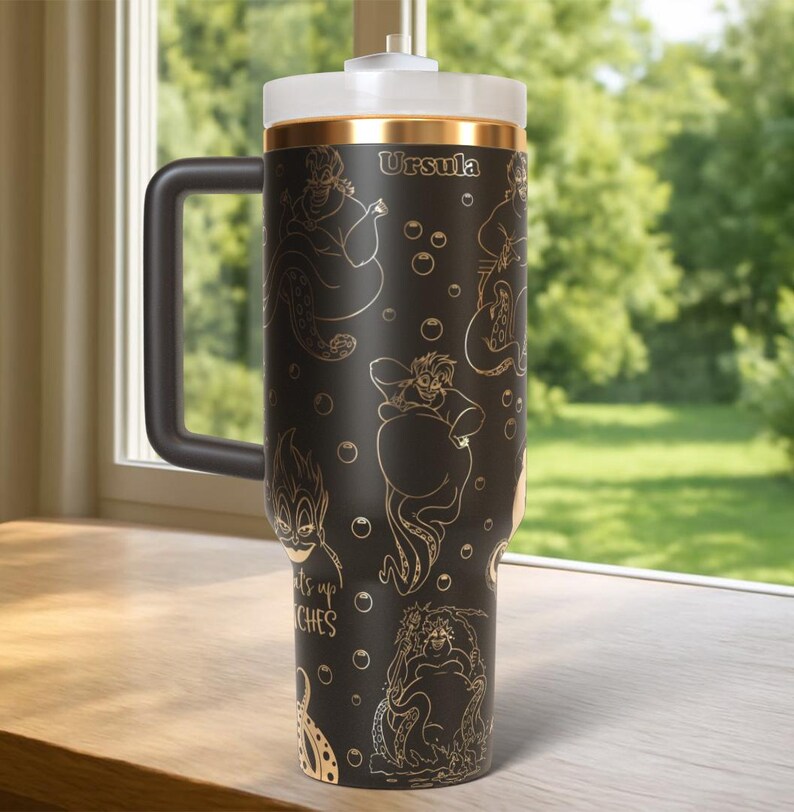 Może przedstawiać: Czarny kubek podr&oacute;żny z białą pokrywką i czarnym uchwytem. Kubek zawiera złote ilustracje Urszuli z Małej Syrenki, z widocznym napisem "Urszula" i frazą "Witches up".