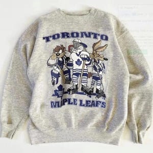 May include: A light grey sweatshirt with the text "TORONTO MAPLE LEAFS" in blue. The graphic shows cartoon characters in hockey uniforms. The sweatshirt has a crew neck and long sleeves.