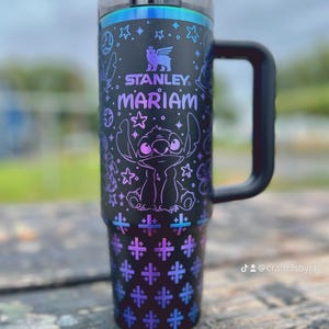 Puede incluir: Vaso Stanley negro con asa, con un dise&ntilde;o morado y azul del personaje Stitch, estrellas y cruces. El nombre "Mariam" est&aacute; impreso en morado. El vaso tiene tapa y pajita negras.