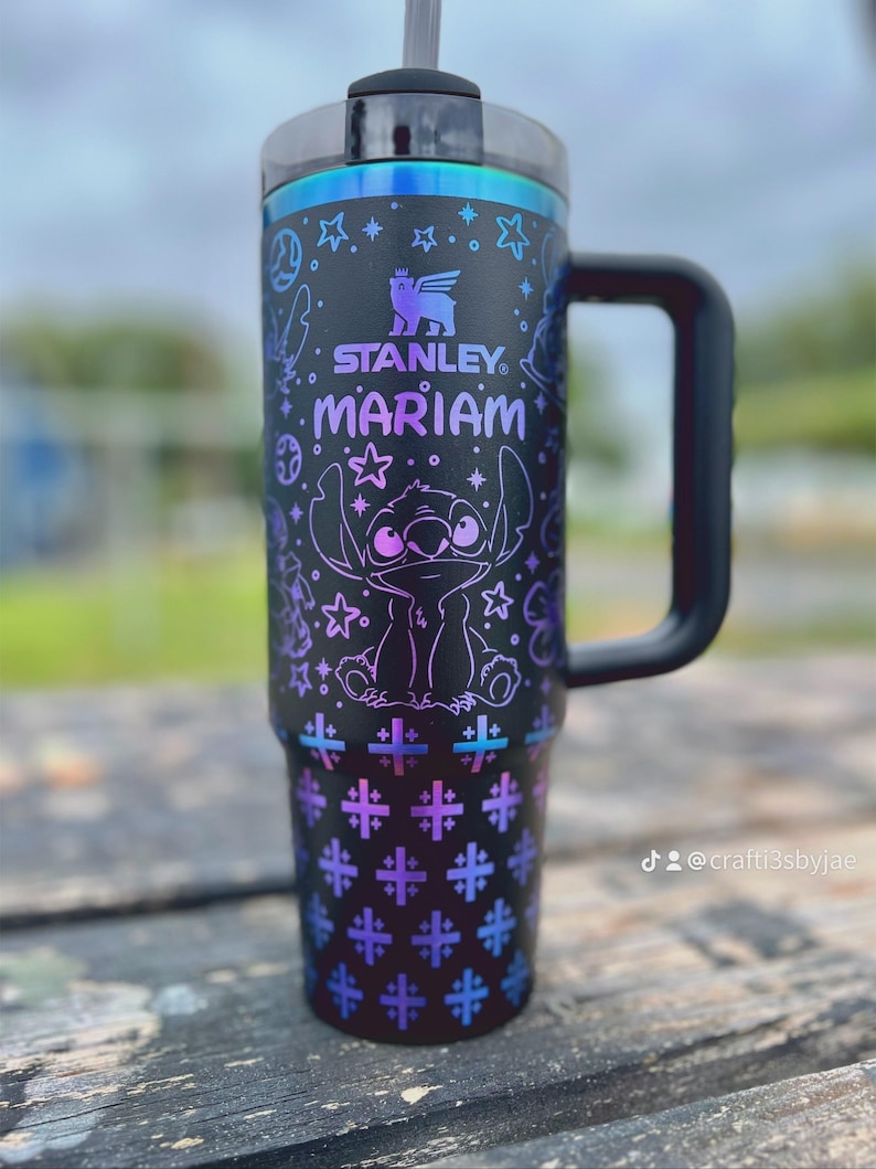 Puede incluir: Vaso Stanley negro con asa, con un dise&ntilde;o morado y azul del personaje Stitch, estrellas y cruces. El nombre "Mariam" est&aacute; impreso en morado. El vaso tiene tapa y pajita negras.