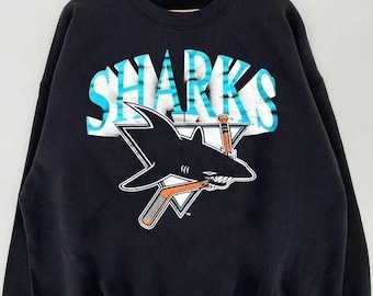 May include: Black sweatshirt featuring the word "SHARKS" in white and teal lettering. A black shark graphic with a hockey stick is centered below the word. The sweatshirt has a crew neck and long sleeves.