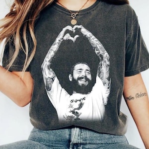 May include: A charcoal gray short-sleeve t-shirt featuring a black and white graphic print of a bearded person with heavily tattooed arms forming a heart shape with their hands above their head. The person wears a white t-shirt with a graphic design.
