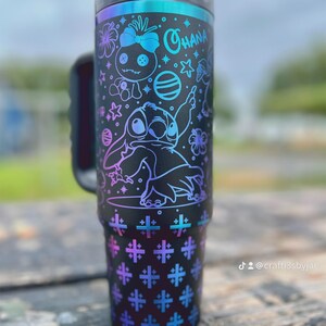 Puede incluir: Vaso negro con asa, con un dise&ntilde;o de Stitch y otros personajes de Lilo & Stitch. El dise&ntilde;o incluye la palabra "Ohana" y varias estrellas y flores. El vaso tiene una tapa negra y una combinaci&oacute;n de colores degradados de azul, morado y verde azulado.