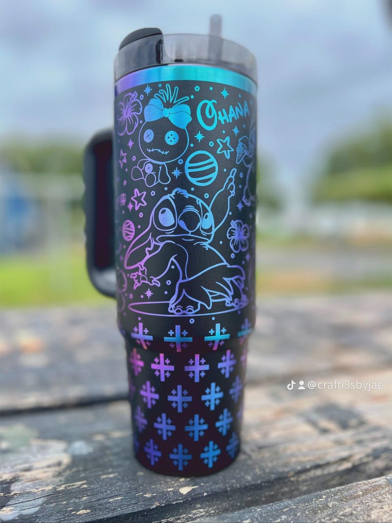 Puede incluir: Vaso negro con asa, con un dise&ntilde;o de Stitch y otros personajes de Lilo & Stitch. El dise&ntilde;o incluye la palabra "Ohana" y varias estrellas y flores. El vaso tiene una tapa negra y una combinaci&oacute;n de colores degradados de azul, morado y verde azulado.