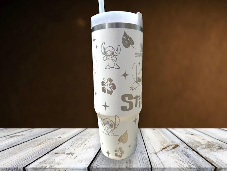 Może przedstawiać: Biały kubek ze srebrną obręczą i pokrywką, z wytrawionymi wzorami postaci Stitcha, gwiazd i tropikalnych kwiat&oacute;w. Kubek zawiera białą słomkę i jest przeznaczony do zimnych napoj&oacute;w.