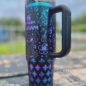 Puede incluir: Vaso t&eacute;rmico negro con asa, con el texto "Stanley" y "Mariam". El vaso est&aacute; decorado con personajes de dibujos animados, estrellas, una guitarra y un estampado floral en tonos morados y azules.