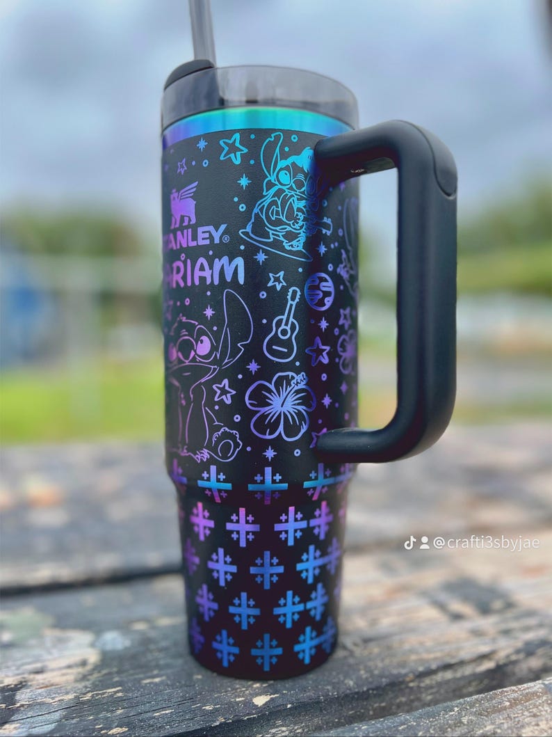 Puede incluir: Vaso t&eacute;rmico negro con asa, con el texto "Stanley" y "Mariam". El vaso est&aacute; decorado con personajes de dibujos animados, estrellas, una guitarra y un estampado floral en tonos morados y azules.