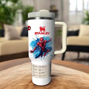 Op de afbeelding: Witte Stanley reismok met een Spider-Man ontwerp. De mok heeft een grijze deksel en handvat, met het Stanley logo zichtbaar. De Spider-Man afbeelding staat tegen een blauwe splash achtergrond.