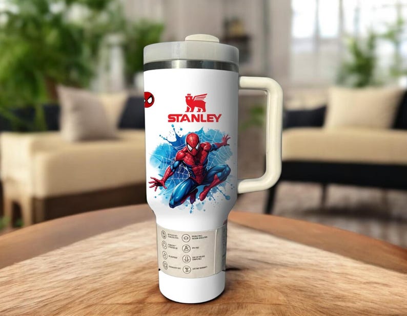 Op de afbeelding: Witte Stanley reismok met een Spider-Man ontwerp. De mok heeft een grijze deksel en handvat, met het Stanley logo zichtbaar. De Spider-Man afbeelding staat tegen een blauwe splash achtergrond.