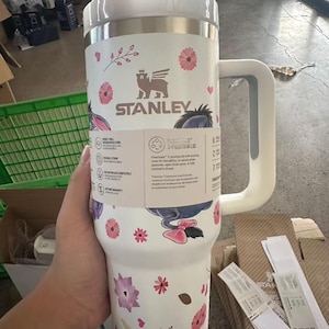 May include: White Stanley travel mug with a floral pattern, including pink and purple flowers and a cartoon character. It has a white handle and lid, with the Stanley logo. Designed for cold drinks, it is suitable for carrying around.