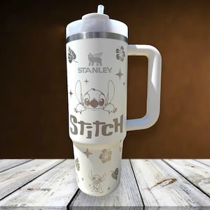Może przedstawiać: Biały kubek Stanley z uchwytem, ​​z motywem postaci Stitch i napisem "Stitch". Kubek ma r&oacute;wnież kwiatowe i gwiazdowe wzory oraz słomkę. Widoczne jest logo Stanley.