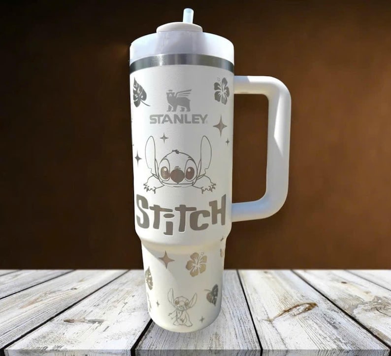 Może przedstawiać: Biały kubek Stanley z uchwytem, ​​z motywem postaci Stitch i napisem "Stitch". Kubek ma r&oacute;wnież kwiatowe i gwiazdowe wzory oraz słomkę. Widoczne jest logo Stanley.