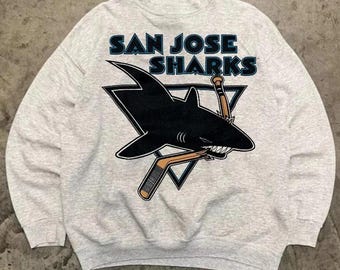 May include: Light grey sweatshirt featuring the San Jose Sharks logo. The logo includes a black shark biting a hockey stick and a hockey puck, set against a black triangle. The words "SAN JOSE SHARKS" are printed above the logo in blue.
