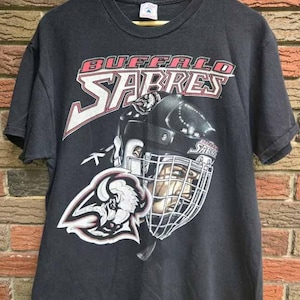 May include: Black t-shirt featuring the Buffalo Sabres logo and team name in red and white. The graphic includes a hockey player's helmet and the team's logo, a buffalo head. The shirt is hanging on a wooden hanger.