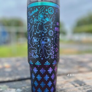 Puede incluir: Vaso negro con tapa y pajita, con un dise&ntilde;o de Stitch de Lilo & Stitch. El dise&ntilde;o incluye la palabra "Ohana" y varias estrellas y formas en azul y morado. La parte inferior tiene un patr&oacute;n repetido de cruces azules y moradas.
